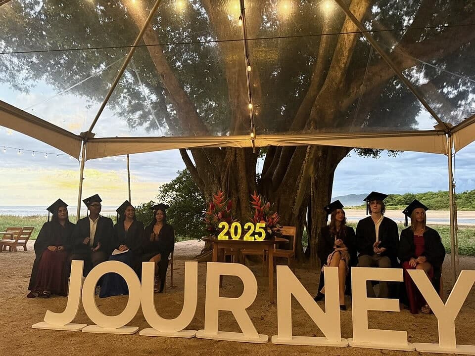 Journey 2025 graduates in caps and gowns under the guanacaste tree at the beach