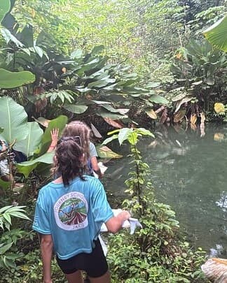 Jacó middle-school student in a Journey teal t-shirt walking along the Las Monas River, dense rainforest around them