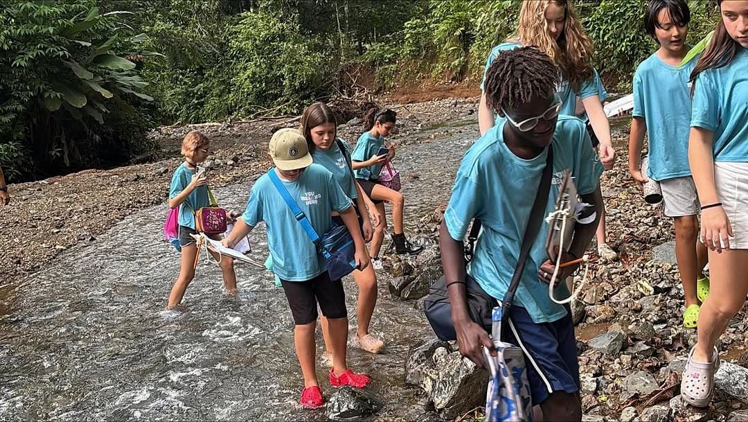 Jacó middle-school students in teal Journey t-shirts wading across rocks on the Las Monas River during an interdisciplinary field trip
