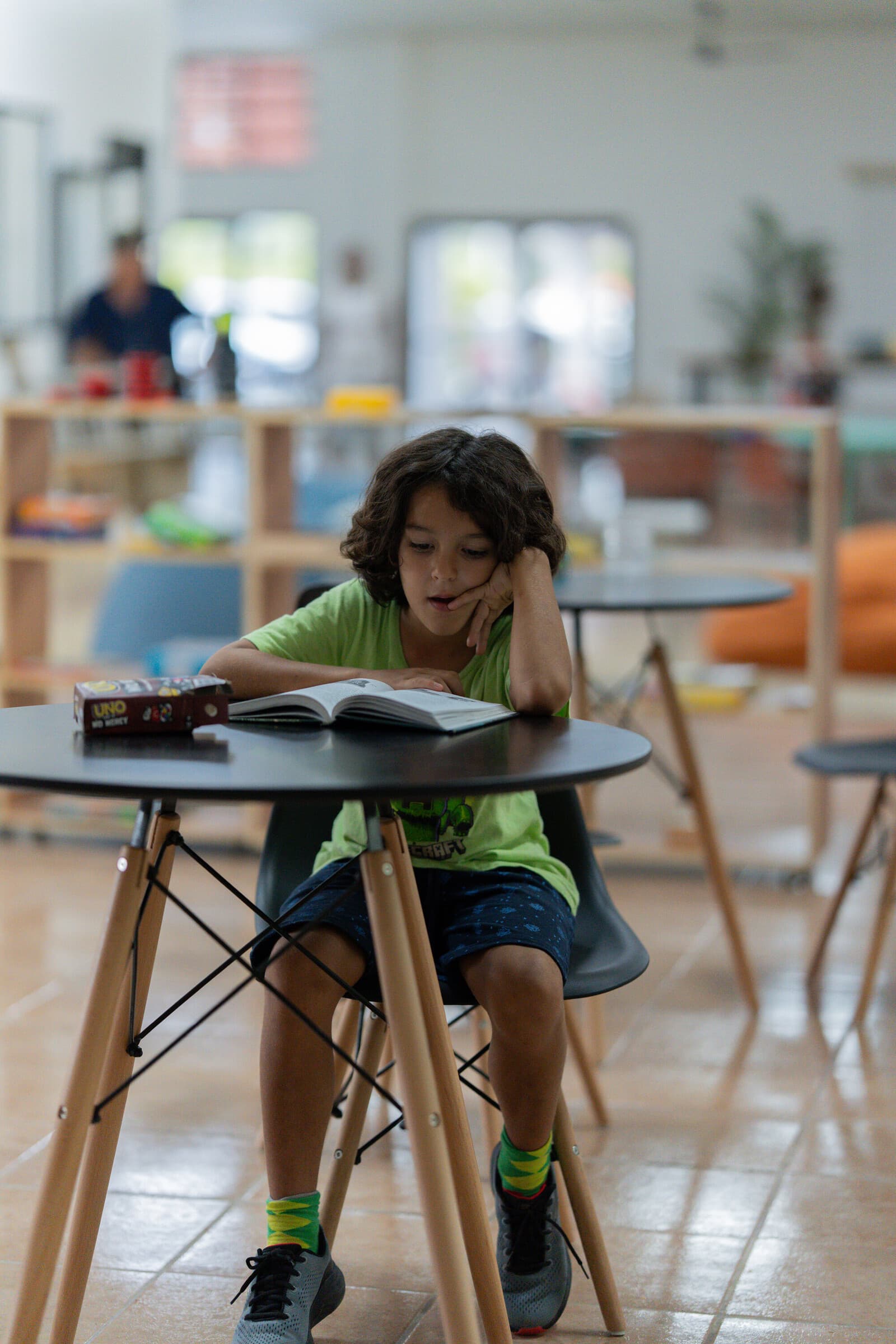 A Journey student in a green Jacó t-shirt absorbed in a book at a small Eames-style table inside the library