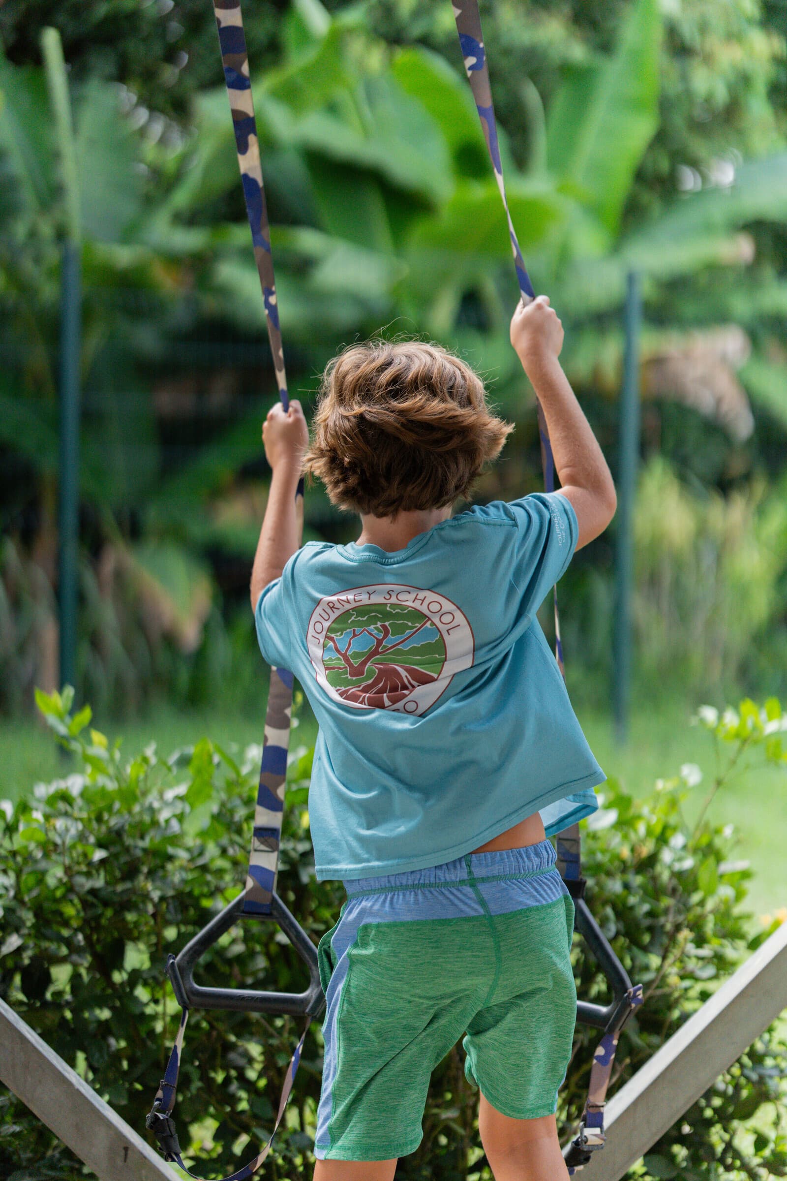 Student from behind in a teal Journey t-shirt swinging on a camo strap-swing in the Jacó rainforest garden