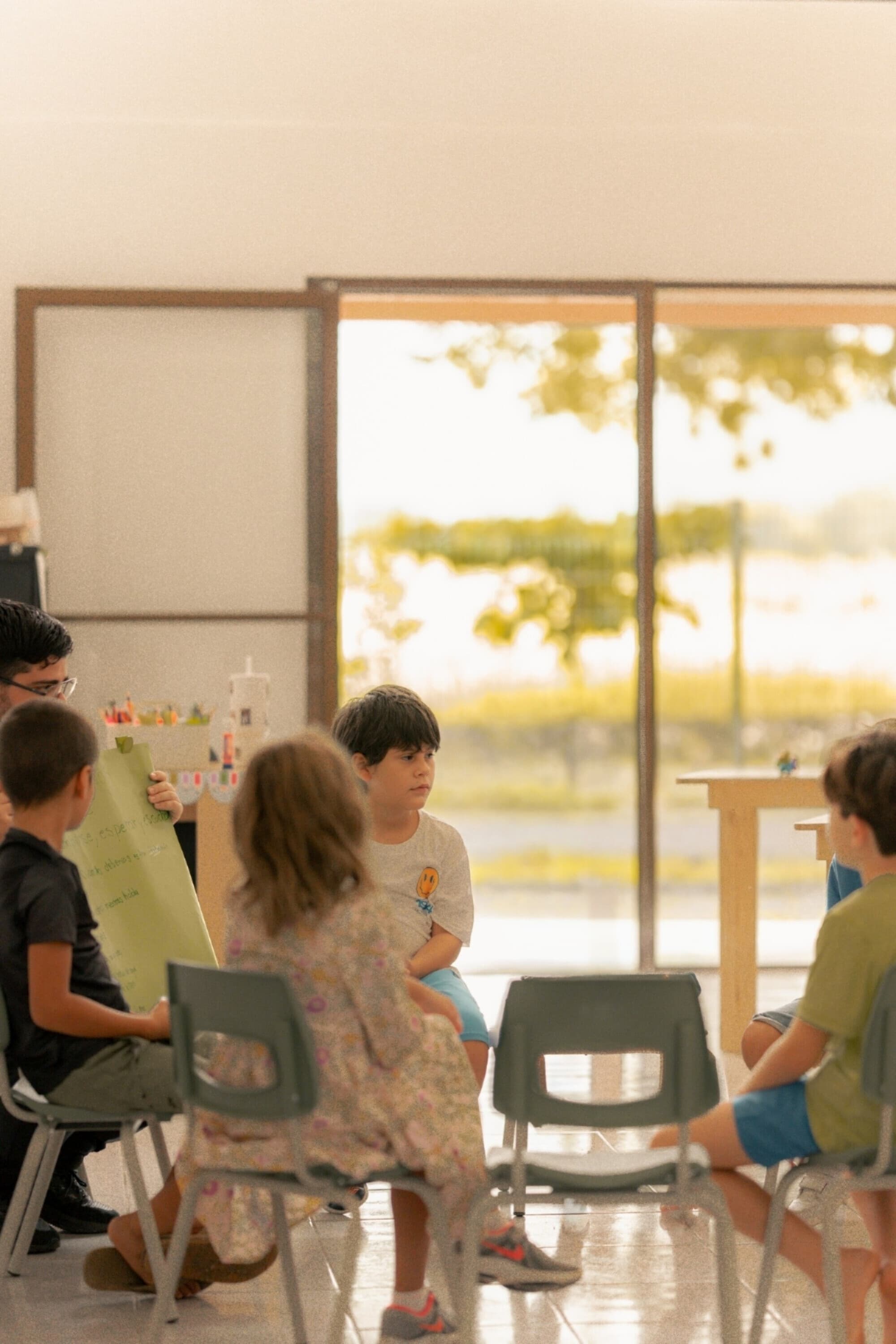 Reggio-style class circle at the Jacó campus — kids around a teacher with a chart-paper easel, golden-hour light