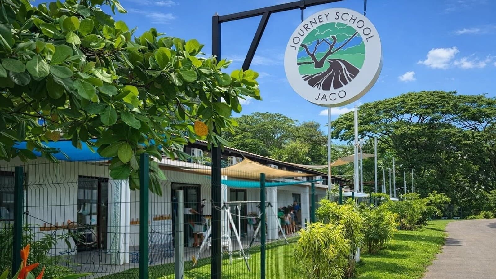 Ground-level view of the JOURNEY SCHOOL JACO entrance sign with the campus building and lush tropical foliage